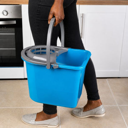A person in black pants and gray slip-on shoes holds a bright blue plastic cleaning bucket with a gray handle and wringer attachment in a kitchen setting.