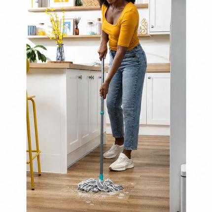 A woman in a yellow top and blue jeans mops a wooden floor in a bright white kitchen with light wood countertops.