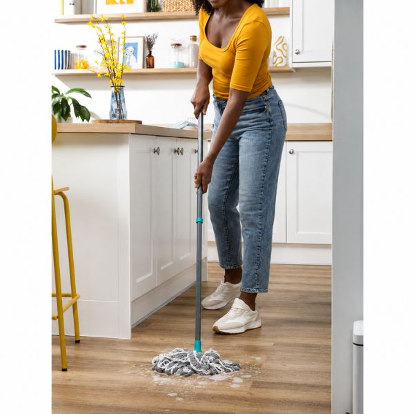 A woman in a yellow top and blue jeans mops a wooden floor in a bright white kitchen with light wood countertops.