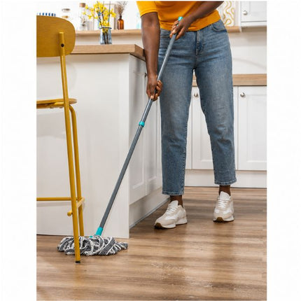 A person mops a wooden floor in a bright kitchen, holding a gray and turquoise-handled microfiber mop. The person is wearing blue jeans, a yellow top, and white sneakers.