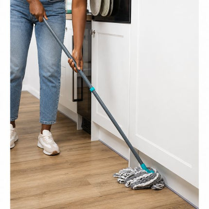 A person mops a light wood floor in a kitchen, holding a gray-handled microfiber mop with turquoise accents near the base of a white cabinet.