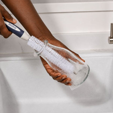 A person holds a glass jar with a white and navy scrubber brush inside a white sink.