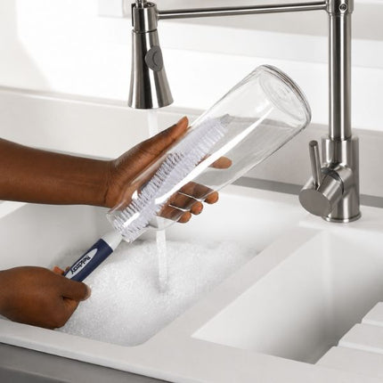 A person uses a Kleanode glass bottle cleaning brush with white bristles and a blue handle to clean a clear glass bottle under a stainless steel kitchen faucet running water in a white sink.