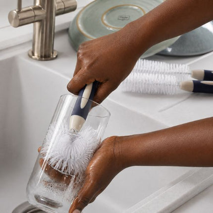 A person's hands wash a clear glass cup with a navy blue and white scrubber brush in a white sink, with more matching brushes visible in the background.