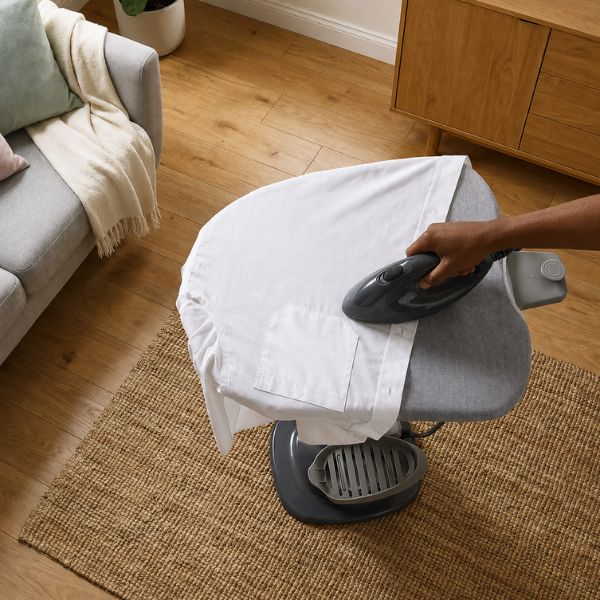 A person uses a dark gray steam iron to iron a white shirt on a gray rolling iron stand in a living room setting with wooden floors and a sofa.
