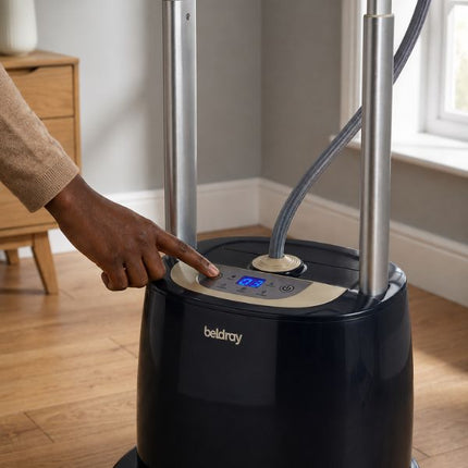 A person's hand pressing the control panel of a black Beldray steam ironing machine in a modern room with wooden floors and gray walls.