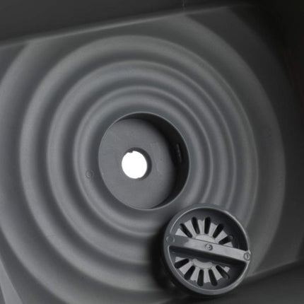 Close-up of a dark gray circular drain or filter component with concentric ring grooves and a small vented cover, shown against a matching dark background.
