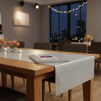 Caroline 90 cm silver square paper table cover draped over a wooden dining table with packaged covers, flowers and candles in a room prepared for hosting.