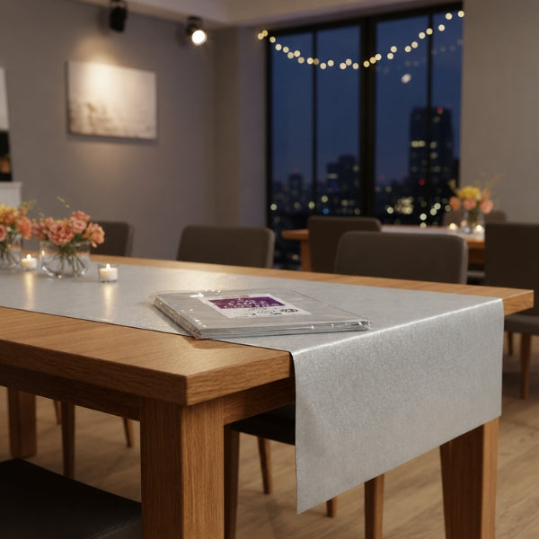 Caroline 90 cm silver square paper table cover draped over a wooden dining table with packaged covers, flowers and candles in a room prepared for hosting.