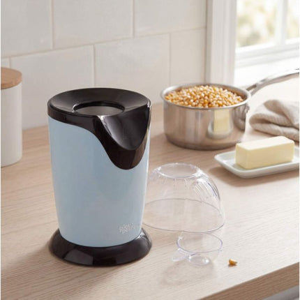 A light blue and black popcorn maker sits on a wooden kitchen counter next to a pot of popcorn kernels, butter, and clear plastic lids.