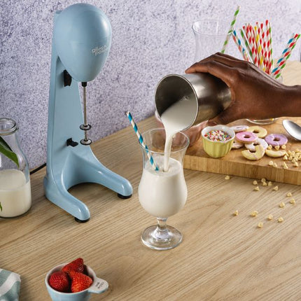 A light blue countertop electric milkshake maker stands on a wooden table next to a hand pouring milkshake into a glass with a striped straw. The scene includes donuts, cereal, additional straws, strawberries, and a bottle of milk in a kitchen-style setting.