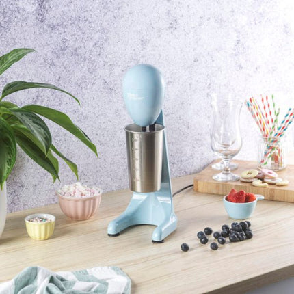 A light blue electric milkshake maker machine with a stainless steel cup, set on a light wood countertop in a kitchen-like setting with a plant, bowls of fruit and whipped cream, and a glass.