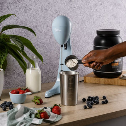 A light blue countertop milkshake maker machine is shown with a hand holding a scoop of powder above a metal measuring cup. A large black protein powder tub, strawberries, blueberries, a glass bottle of milk, and a potted plant are arranged on a wooden table against a textured light gray wall.