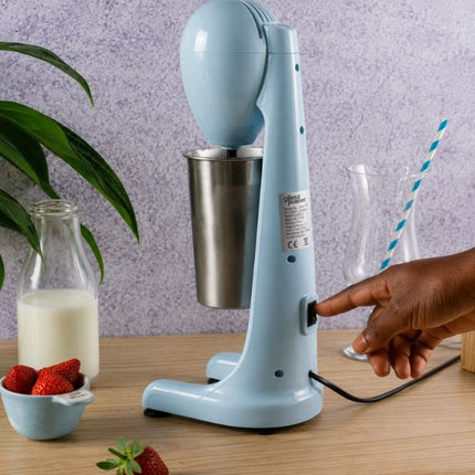 A light blue electric milkshake mixer with a stainless steel cup, shown on a wooden table with a hand turning its switch. The scene includes a glass bottle of milk, strawberries, and a blue-and-white striped straw against a textured purple wall.