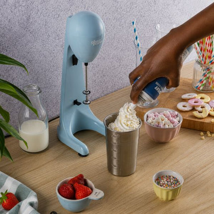 A light blue countertop whipped milkshake maker sits on a wooden table, with a hand adding whipped cream from a canister into a metal cup. Bowls of strawberries, marshmallows, sprinkles, donuts, and striped straws are arranged around it against a textured light purple wall.