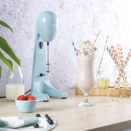 A light blue countertop milkshake mixer stands beside a tall glass filled with a pink milkshake topped with sprinkles and a straw on a wooden table. A small bowl of strawberries, a glass bottle of milk, and additional straws are arranged around it in a bright kitchen-like setting.