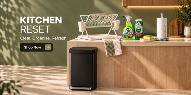 A black rectangular kitchen trash can with a lid stands on a light wood floor beside a wooden kitchen counter. The scene includes a white dish rack, cleaning spray bottles, a towel, paper towel holder, and a green wall with the text 'KITCHEN RESET Clear.