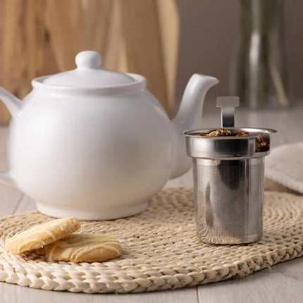 A stainless steel mesh infuser basket with a fold-over handle, sitting next to a white teapot