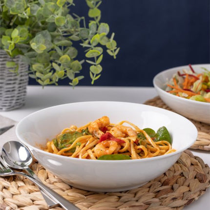 A white ceramic bowl filled with shrimp and noodle pasta, placed on a woven placemat on a table with a spoon, another bowl of salad, and a potted plant in the background.