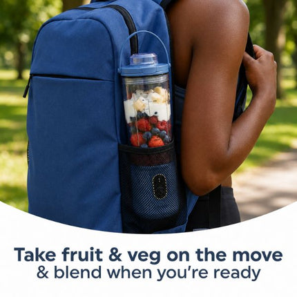 A blue backpack worn outdoors by a person, shown from the back with a clear blending bottle attached in the side pocket. The bottle contains mixed fruit and vegetables, and text at the bottom reads, 'Take fruit & veg on the move & blend when you're ready.