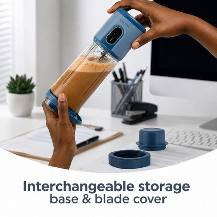A portable blender bottle with a blue storage base and matching blue blade cover, shown being held by two hands at a desk. The bottle is partially filled with a brown beverage.