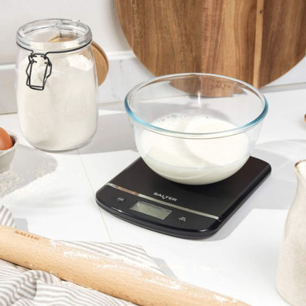 A black digital kitchen scale with a clear glass bowl of milk on top, set on a white countertop with a flour jar, rolling pin, and wooden cutting board in the background.