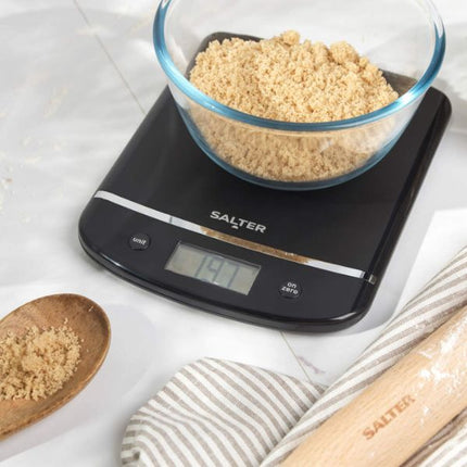 A black digital kitchen scale with the brand name SALTER, shown with a glass bowl filled with coarse beige crumbs on top. The scale is photographed on a light marble-like surface with a wooden spoon of crumbs, a striped cloth, and a wooden rolling pin nearby.