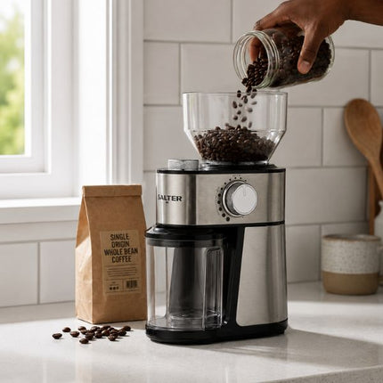 A countertop coffee grinder with a clear hopper being filled with coffee beans from a glass jar, with a person's hand pouring the beans. A paper bag of single-origin whole bean coffee stands nearby on a white kitchen counter.