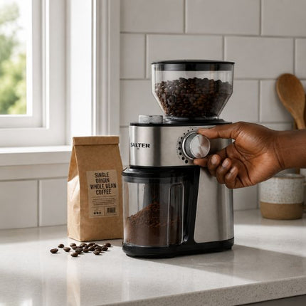 A hand is turning the dial of a Salter electric coffee grinder on a kitchen counter, with a bag of whole bean coffee and loose beans nearby.