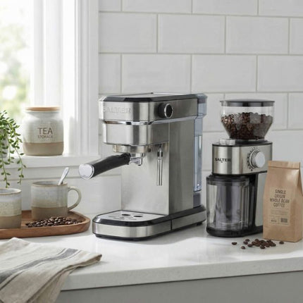 A stainless steel Salter espresso machine sits on a white kitchen countertop next to a matching coffee grinder and a bag of coffee beans, with a tiled backsplash and window in the background.