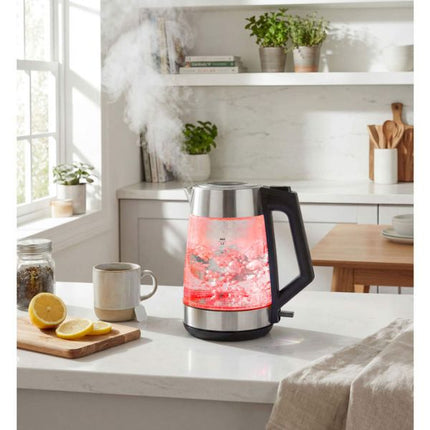 A glass electric kettle with a black handle and base sits on a white kitchen counter, glowing with red light from the water inside. Steam rises from a mug nearby, and a bright kitchen with shelves, plants, and a window is in the background.