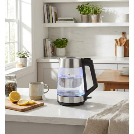 A modern electric kettle with a clear glass body, stainless steel base, and black handle, sitting on a white kitchen countertop next to a mug and lemon slices.