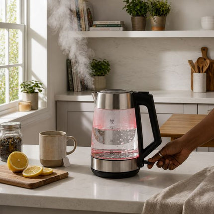 A glass electric kettle with a black handle and base sits on a white kitchen counter, with a person's hand turning the dial. Steam rises from the water, and a mug, lemon slices, and kitchen shelves are visible in the background.