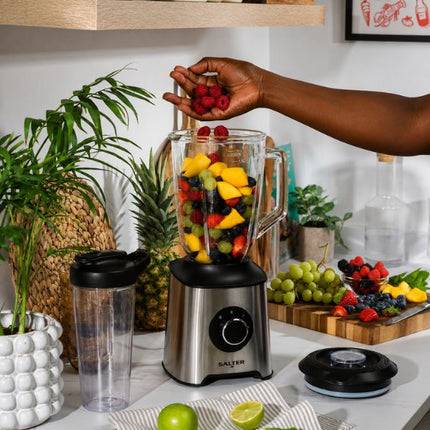 A countertop scene showing a SALTER blender with a clear pitcher filled with mixed fruit, a person's hand adding raspberries, a clear travel cup, and other fruits and kitchen items on a white counter.