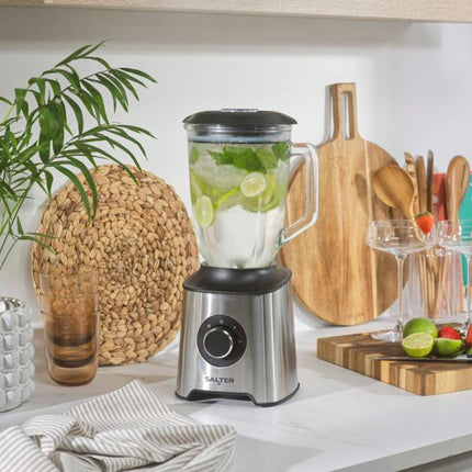 A Salter countertop blender with a stainless steel base and black lid, filled with a green and white beverage, set on a white kitchen counter with cutting boards, glasses, and limes in the background.