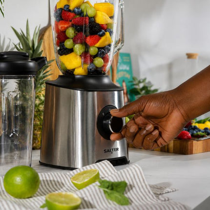 A stainless steel SALTER blender with a clear pitcher filled with mixed fruit, shown on a kitchen counter with a person's hand turning the dial. Limes, mint, and other fruits and vegetables are visible in a bright kitchen setting.