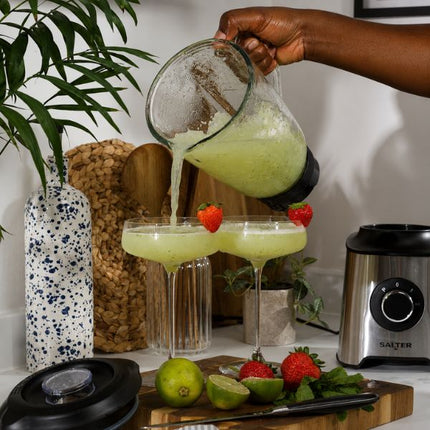 A hand pours pale yellow-green juice from a blender pitcher into two stemmed glasses garnished with strawberries, set on a kitchen counter with limes, strawberries, a blender base, and decorative props in the background.