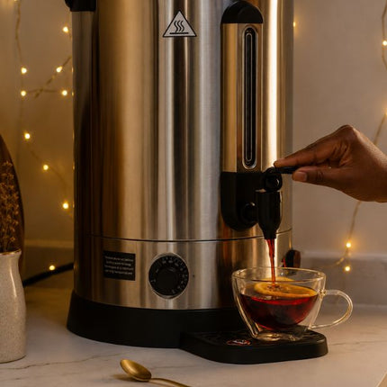 A stainless steel and black commercial coffee dispenser with a hand turning the spout on, pouring hot coffee into a clear glass mug on a marble countertop with warm string lights in the background.