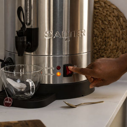 A stainless steel Salter tea maker on a white countertop with a hand turning the power switch. A clear glass teacup, a tea bag, and a spoon are placed nearby, with a woven basket visible in the background.