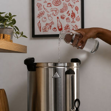 A stainless steel commercial coffee maker with black lids, shown with a person pouring water from a glass pitcher into it. The background features a light gray wall with a framed red food-themed print and a wooden shelf with a plant.