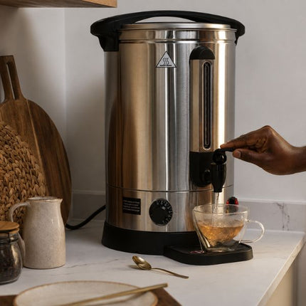 A stainless steel hot water dispenser with a black handle and base, shown dispensing hot water into a clear glass cup on a kitchen counter. A person's hand is visible turning the water level knob.