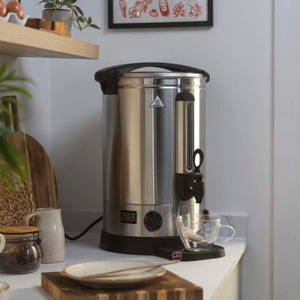 A stainless steel commercial coffee or tea dispenser with a black handle and base, sitting on a kitchen counter next to a glass cup.