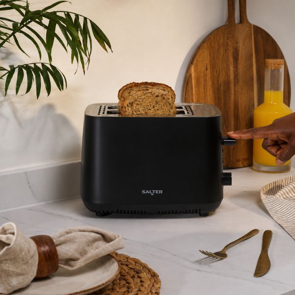 A black Salter toaster with a slice of bread on top, shown on a kitchen counter with a hand pressing the turn knob. A wooden cutting board, a glass bottle of orange juice, and other kitchen items are visible in the background.