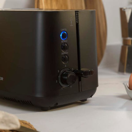 A close-up of a matte black toaster on a white countertop, showing the control panel with a glowing blue power button and rotary dial.