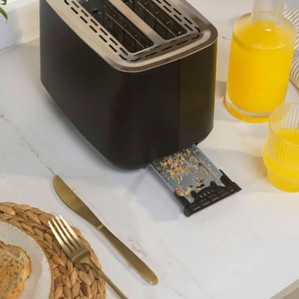 A black and stainless steel toaster sits on a white marble countertop with a bread slice, gold-toned fork, and knife nearby. Two glasses of orange juice are visible in the background, along with a small debris-catching filter hanging from the toaster's slot.