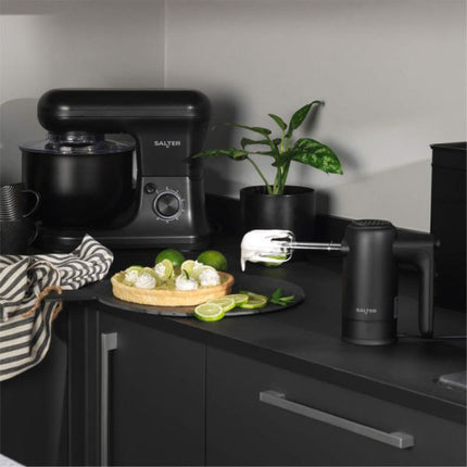 A black SALTEEN countertop mixer and hand mixer set on a dark kitchen counter, with a stand mixer bowl on the left and a handheld attachment on the right. A small potted plant, limes, and a striped cloth are nearby in a modern kitchen setting.