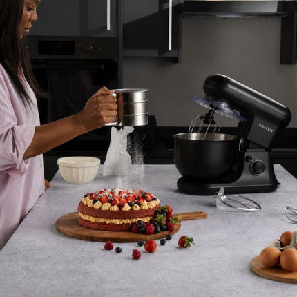 A woman in a pink shirt sifts flour into a bowl of a black SALTER stand mixer on a kitchen counter, with a decorated cake and eggs nearby.