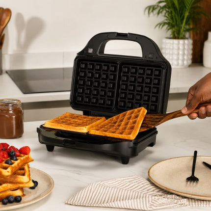 A black electric waffle maker sits on a kitchen counter with two golden-brown waffles inside, being lifted by a wooden spatula held by a person. Plates with waffles and berries, a jar of syrup, and a potted plant are visible in a bright kitchen setting.