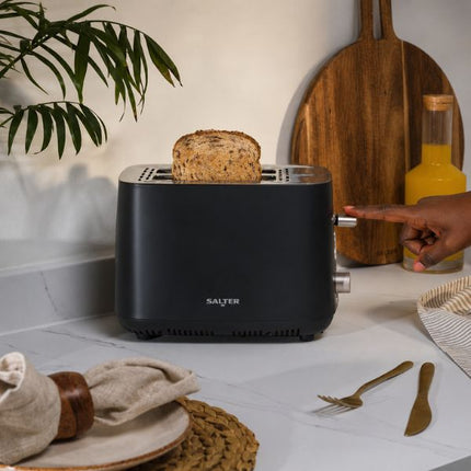 A black Salter toaster with a slice of bread on top, set on a white marble kitchen countertop with a person's hand pointing at the side dial.