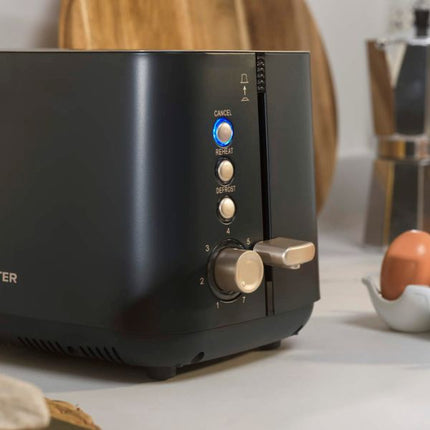 A close-up of a black toaster on a kitchen counter, showing the control buttons and dial, with a wooden cutting board and kitchen items blurred in the background.